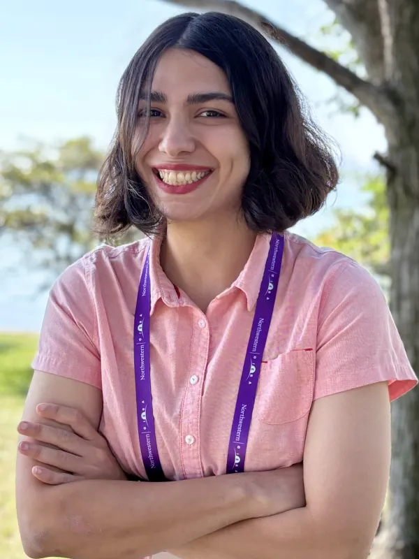 A smiling woman wearing a pink shirt and a purple lanyard with trees in the background